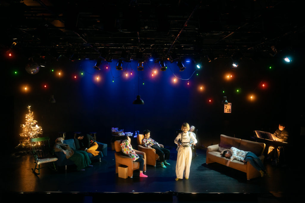 2025年5月「パンとバラで退屈を飾って、わたしが明日も生きていくことを耐える。」舞台公演の写真。クリスマスツリーのある空間で、ソファに座って眠る人々。その中で、白いブラウスに白いズボンの女性（大川翔子）が一人立って、観客に向けて話しかけている。