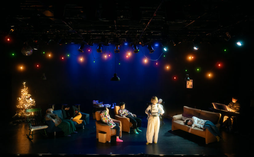 クリスマスツリーのある空間。ソファに座って眠る人々。その中に一人、白いブラウスと白いズボンを着た女性が立って、客席に話しかけている。
